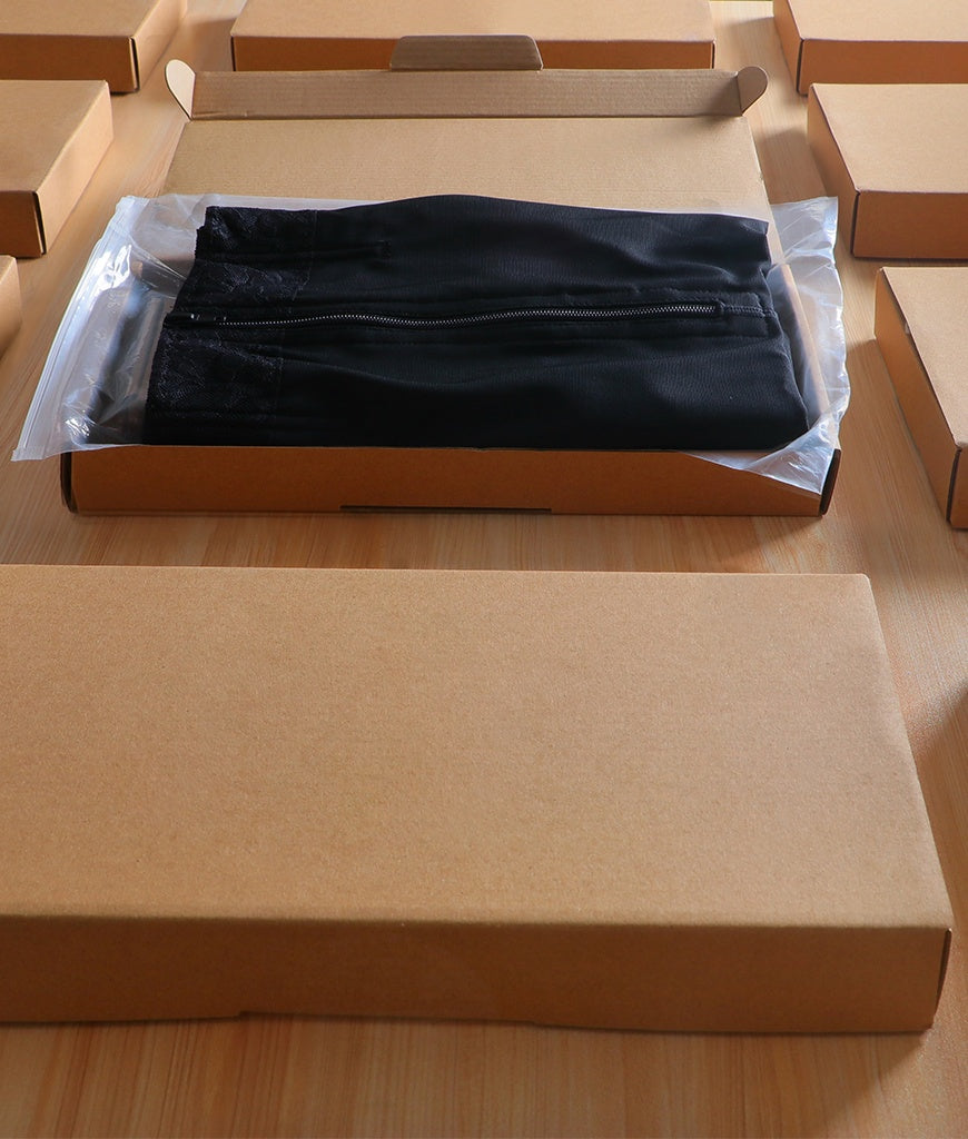 Black women's tummy control body shaper folded in clear plastic packaging inside a brown cardboard box